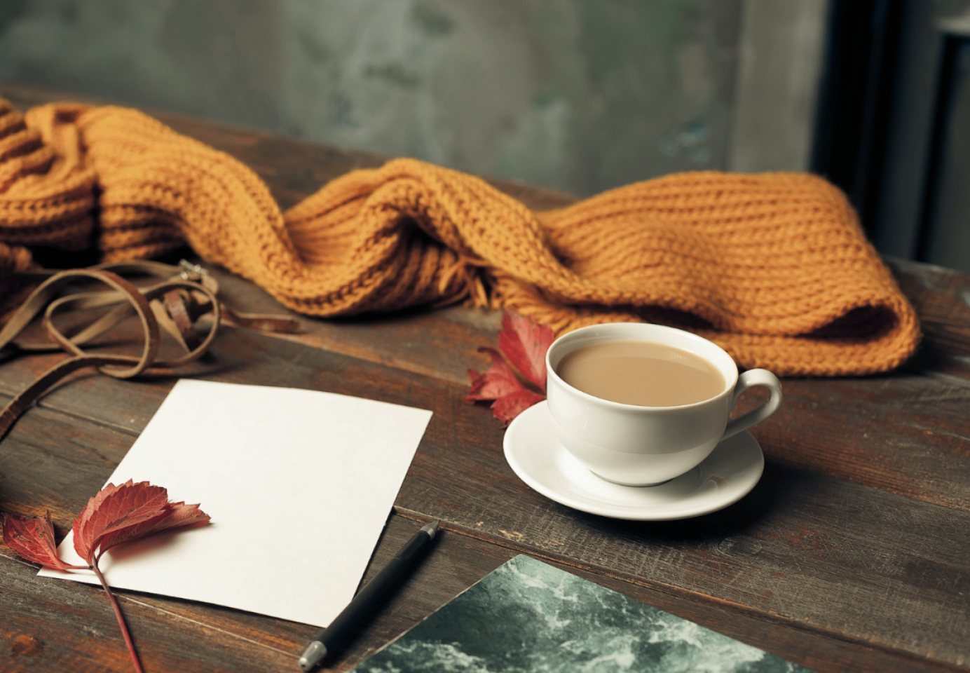 Cup of coffee on a wooden table with a mustard knit scarf, red autumn leaves, and a blank note card, part of a fall self-care routine