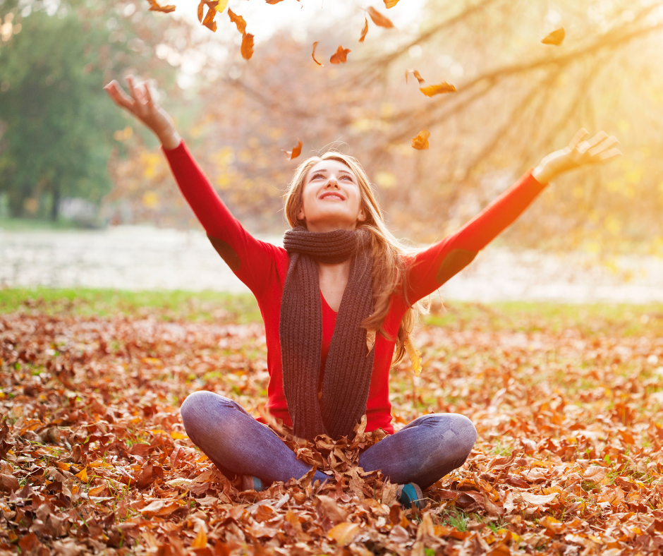 Smiling woman sitting in golden autumn leaves, tossing them into the sunlight as she welcomes fall